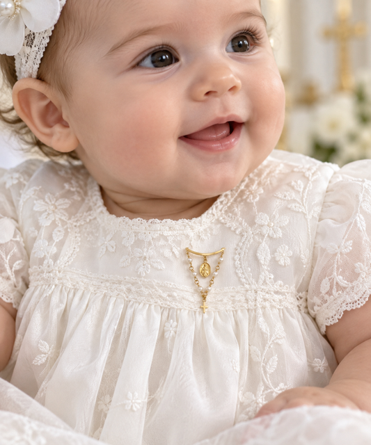 A close-up of a smiling baby wearing a white embroidered lace dress with a gold pearl with a medal and cross baby pin at the neckline and a white floral headband.