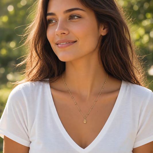 A woman with long brown hair wearing a delicate gold necklace with a small oval pendant, photographed outdoors with a soft green bokeh background.