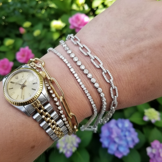 A close-up of a person's wrist wearing multiple bracelets and a watch outdoors, with blurred green foliage and pink and purple flowers in the background. The bracelets include silver and gold-toned chain styles with clear stones, and a two-tone wristwatch with a gold dial.