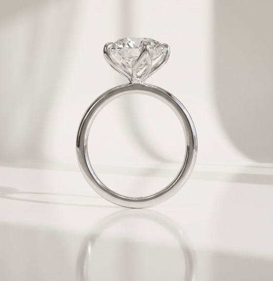 A white gold solitaire engagement ring with a round-shaped diamond gemstone, displayed on a reflective white surface with soft window light shadows.