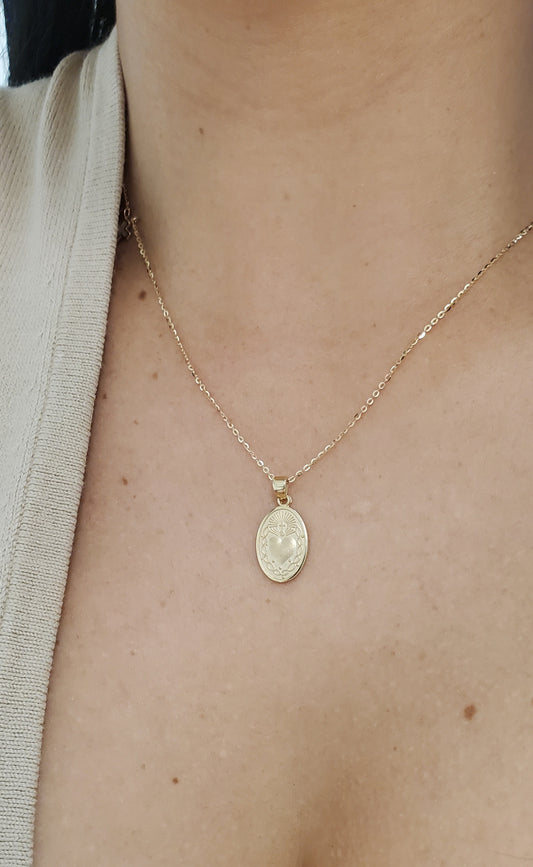 A close-up of a person's neck and chest wearing a delicate gold chain necklace with an oval Sacred Heart of Jesus Medal pendant.