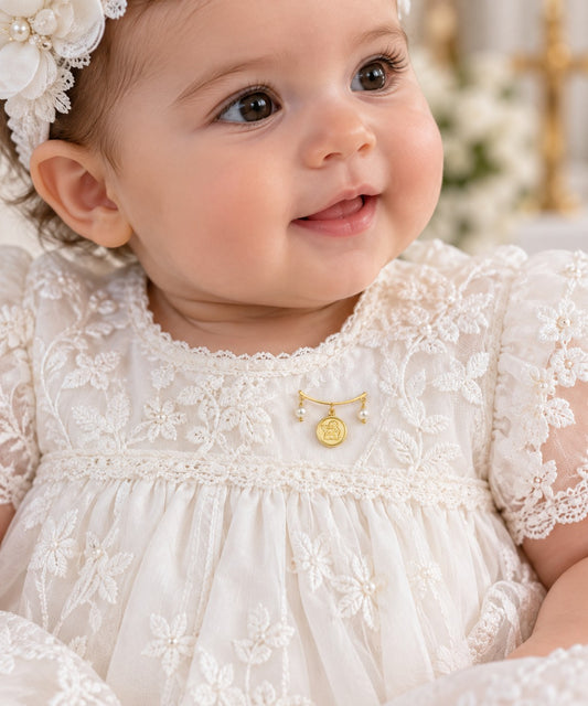 A close-up of a smiling baby wearing a white lace dress with floral embroidery, a matching white flower headband, and a small 14k gold Angel Medal Baby Pin.