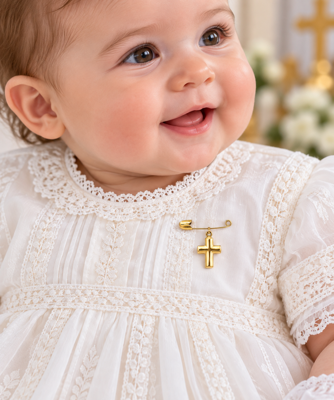 A close-up of a smiling baby wearing a white lace christening dress and a small gold cross pin at the chest, with a softly blurred warm-toned background.