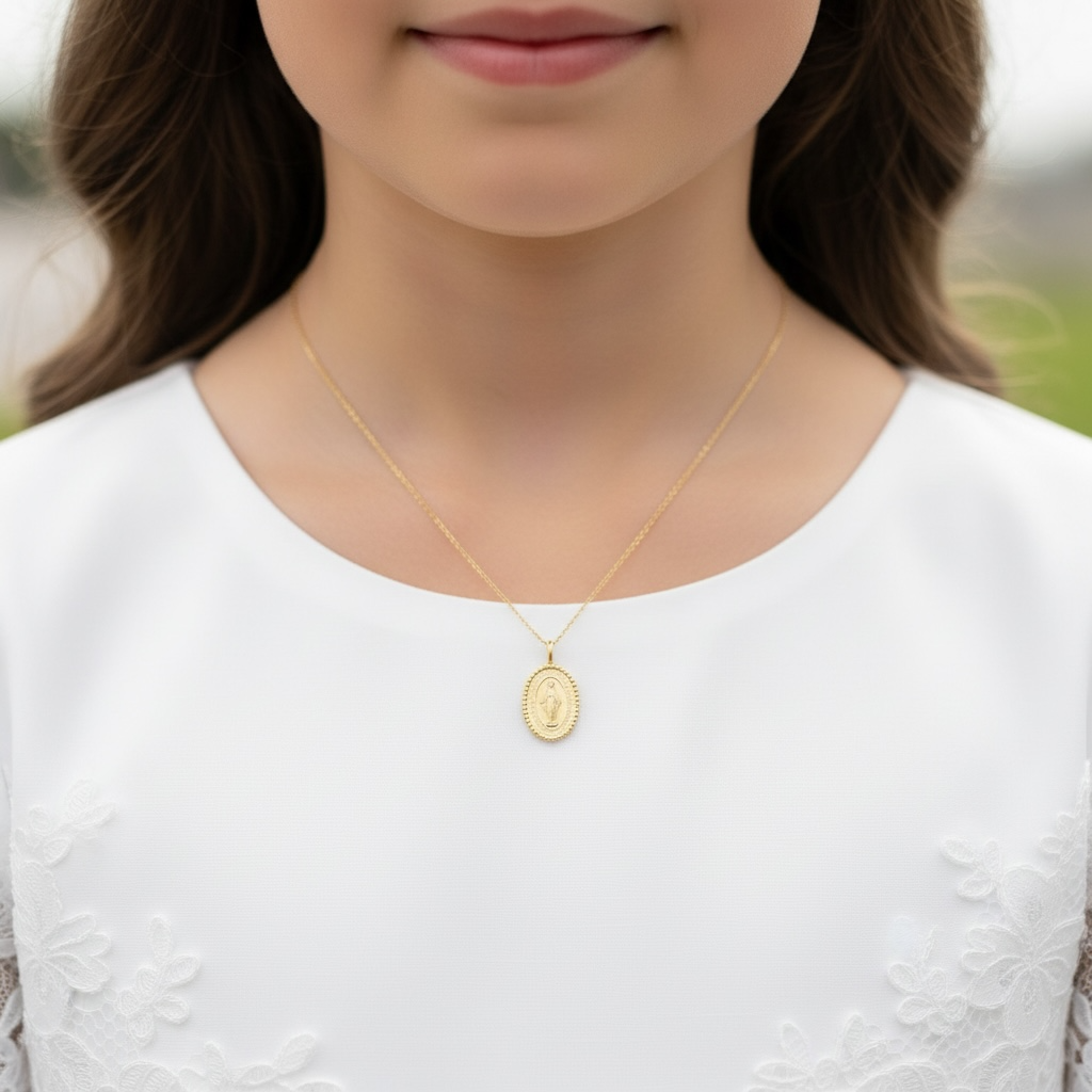 A close-up of a model wearing a delicate gold chain necklace with an oval miraculous medal pendant, styled with a white embroidered top.