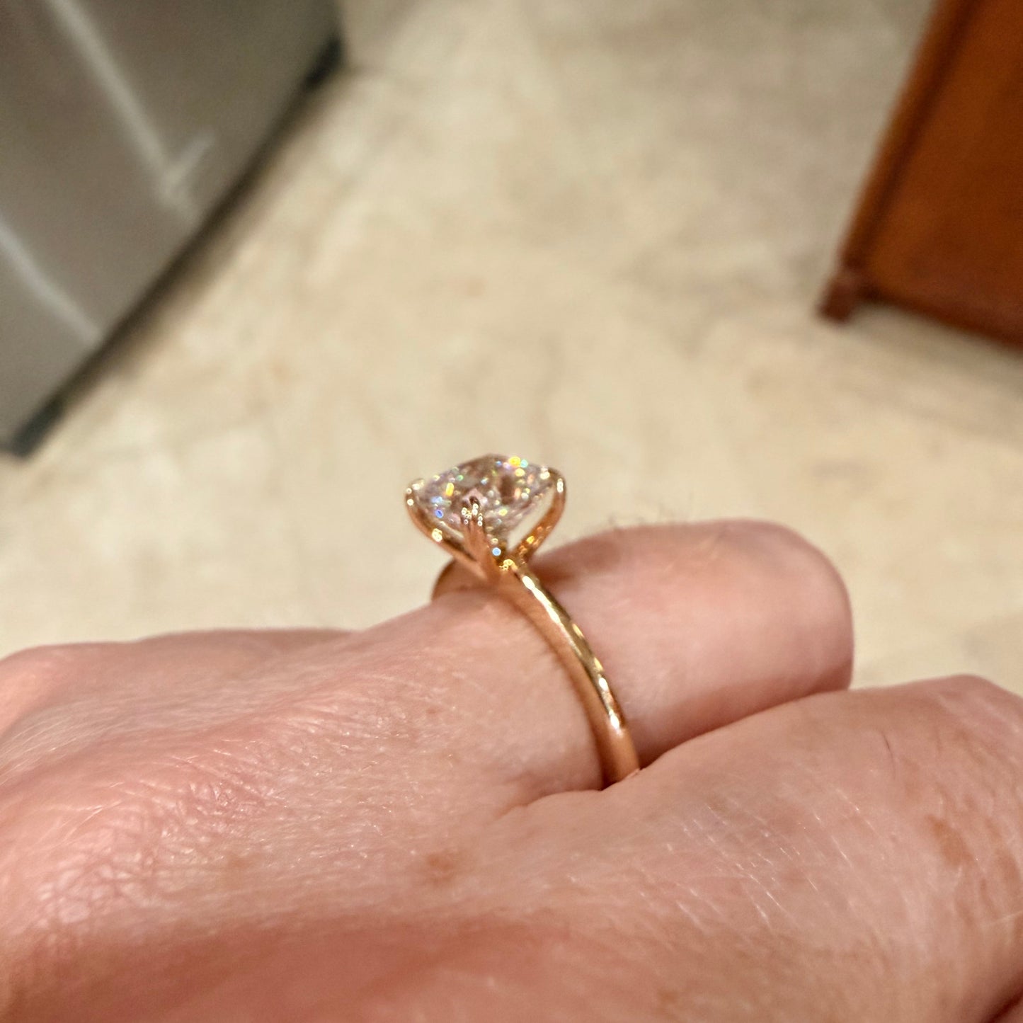 A close-up of a hand wearing a yellow gold solitaire engagement ring with a cushion-shaped diamond gemstone, set against a blurred indoor background.