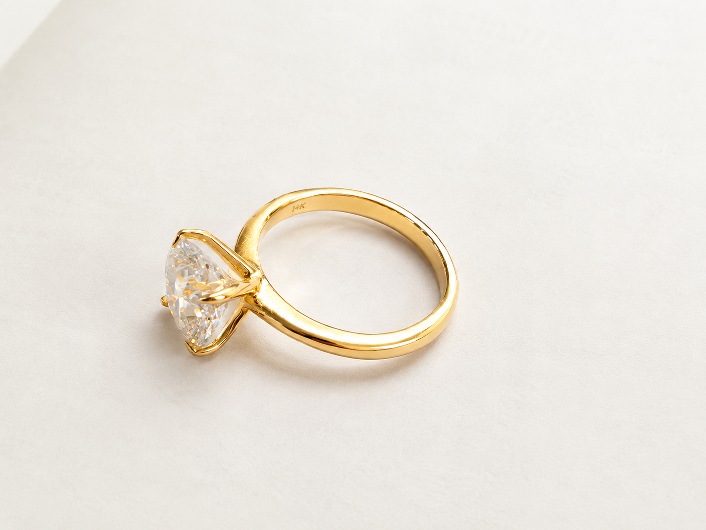 A yellow gold  solitaire engagement ring featuring a large, prong set cushion cut diamond gemstone, photographed on a clean, light off-white background.