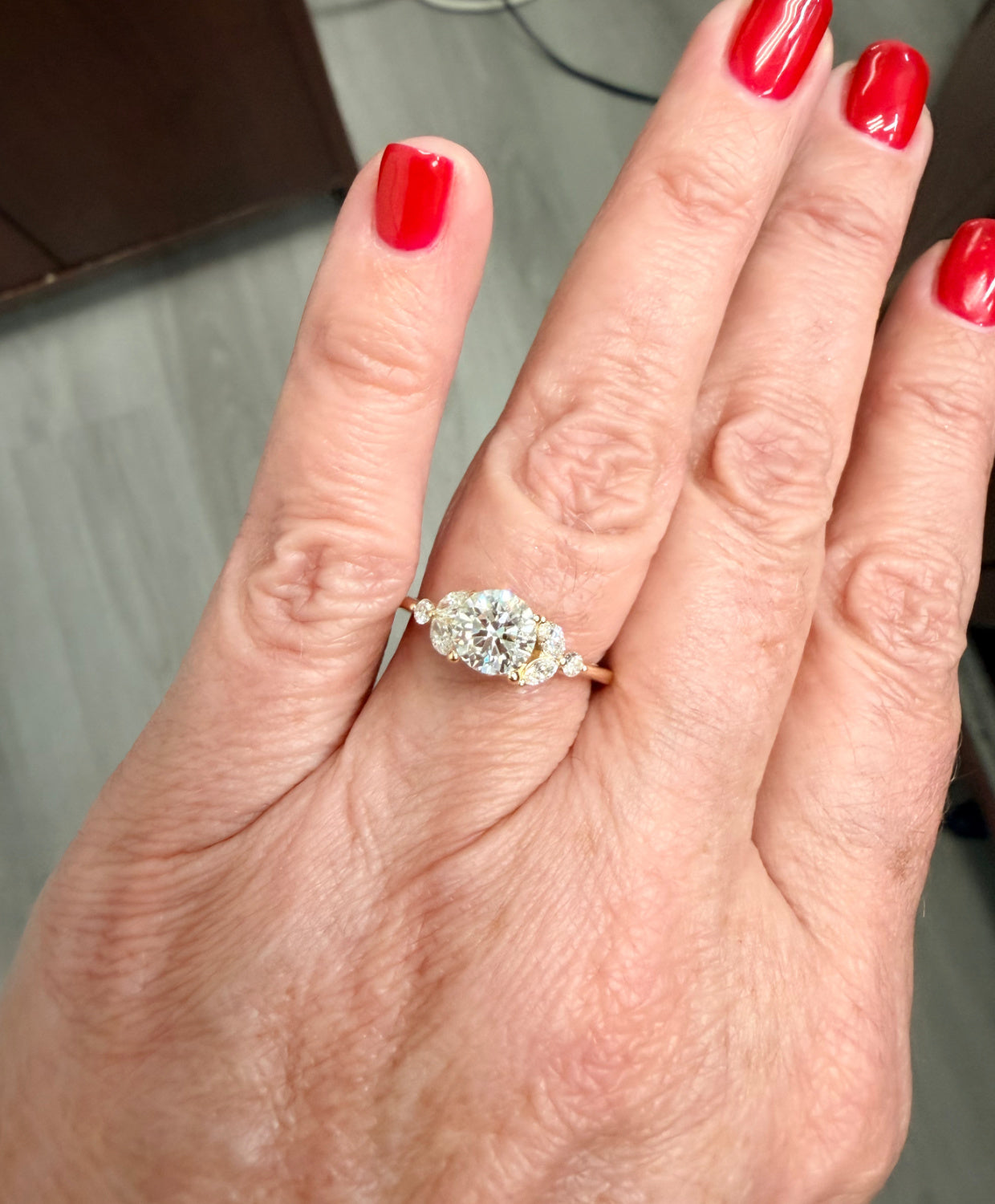 Hand wearing a diamond engagement ring with red nail polish on a neutral background