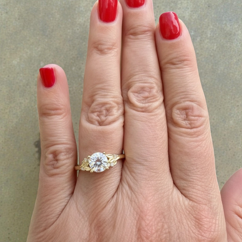Gold engagement ring with a diamond on a hand with red nail polish against a neutral background