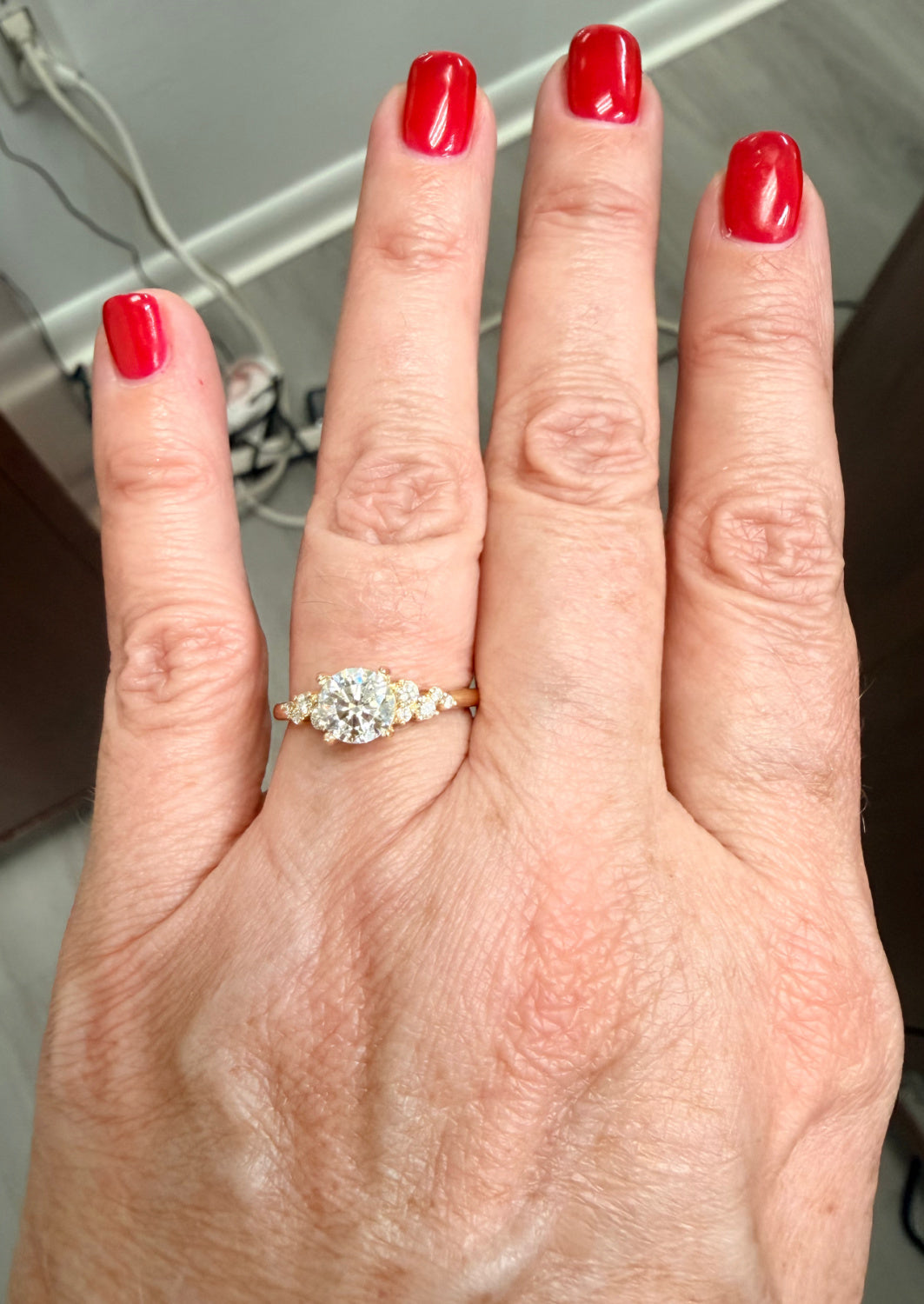 Hand wearing a diamond engagement ring with red nail polish on a neutral background