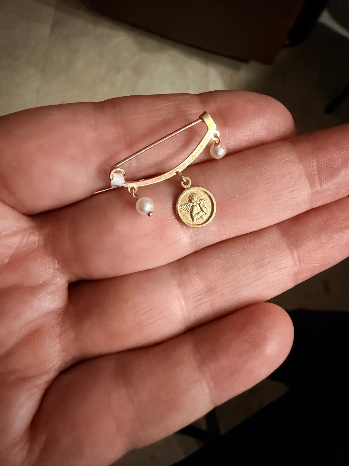 A delicate 14K gold pin brooch with a curved bar, small pearl beads, and a round Angel charm, resting on a person's hand.