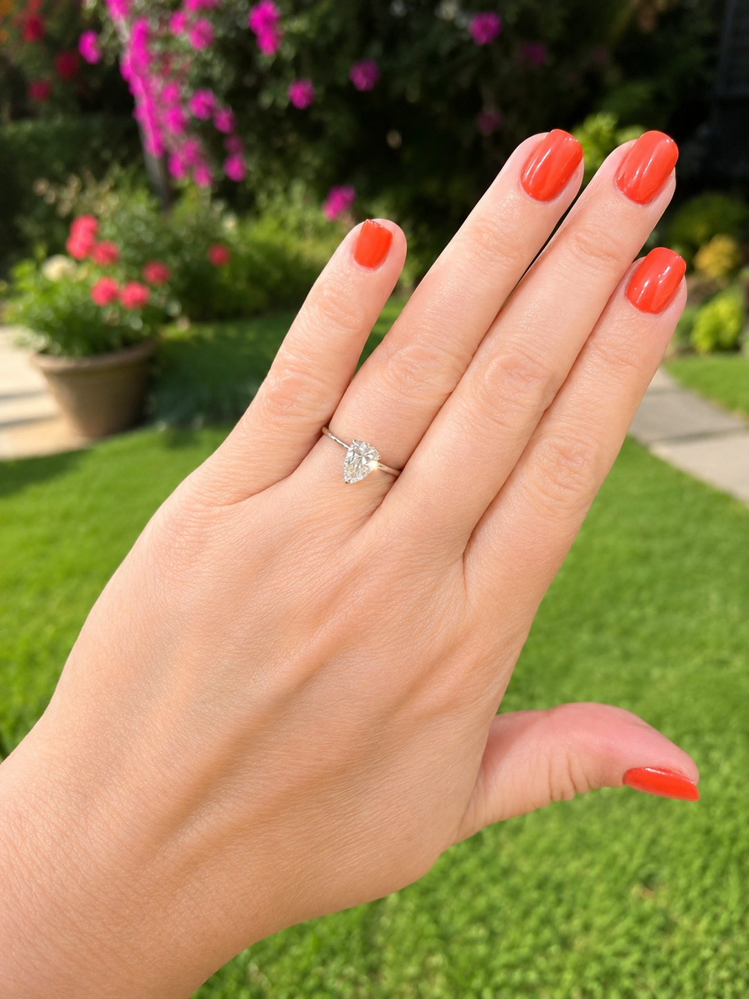A close-up of a hand with bright coral nail polish wearing a white gold engagement ring ring with a Pear-shaped diamond gemstone, set against a blurred green garden background.