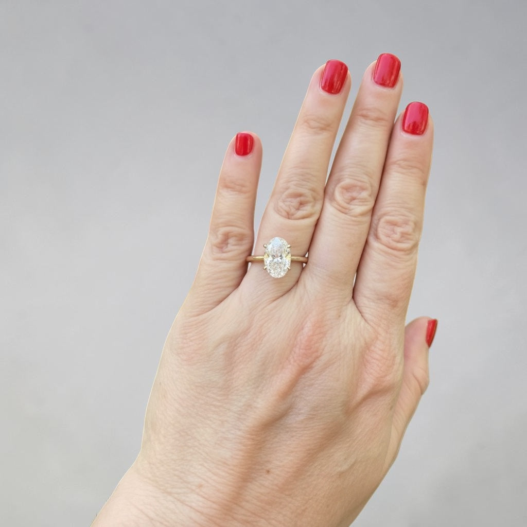 A hand with red-painted nails wearing a yellow gold solitaire engagement ring with a large oval-cut diamond gemstone, set against a plain light gray background.