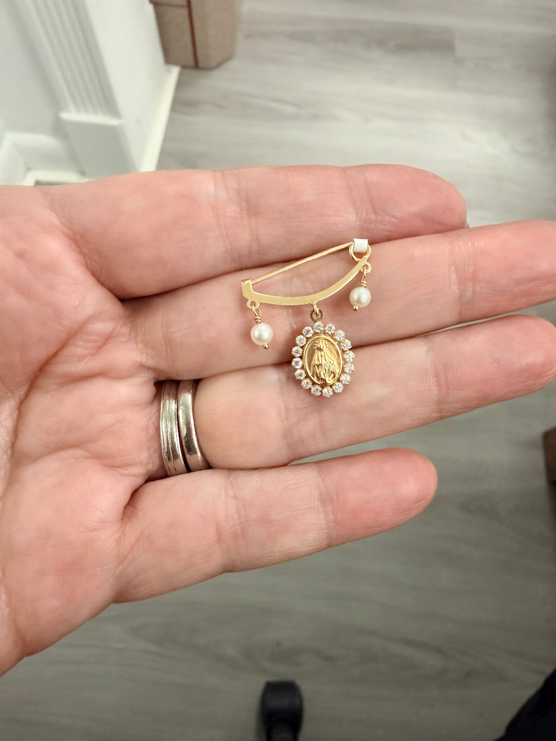 A gold and pearl religious brooch featuring a central oval medallion of the Virgin Mary, held on a person's hand. The brooch has a gold safety pin base with two small dangling pearls on either side of the medallion.
