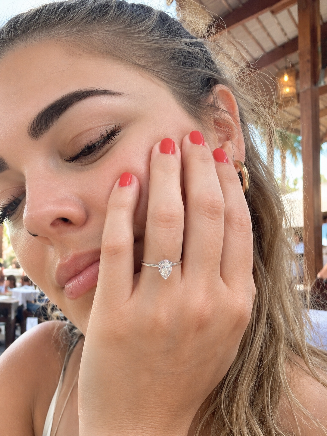 A close-up lifestyle shot of a woman wearing a white gold pear-shaped diamond engagement ring on her finger, with her hand resting on her cheek. She has coral-pink nail polish, and the background shows a warm, rustic outdoor setting.