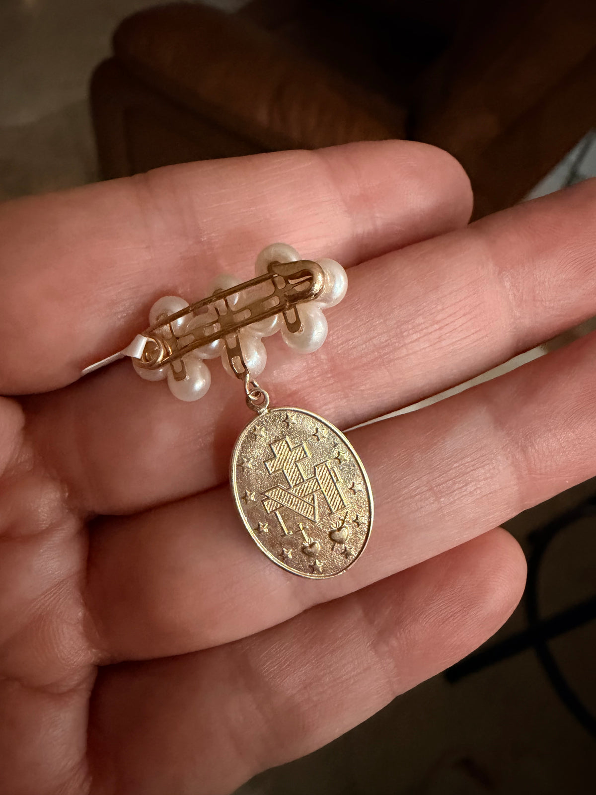 A gold-toned back view of the Miraculous Medal with a pearl-beaded pin and an oval pendant featuring a religious emblem, held in a person's hand.