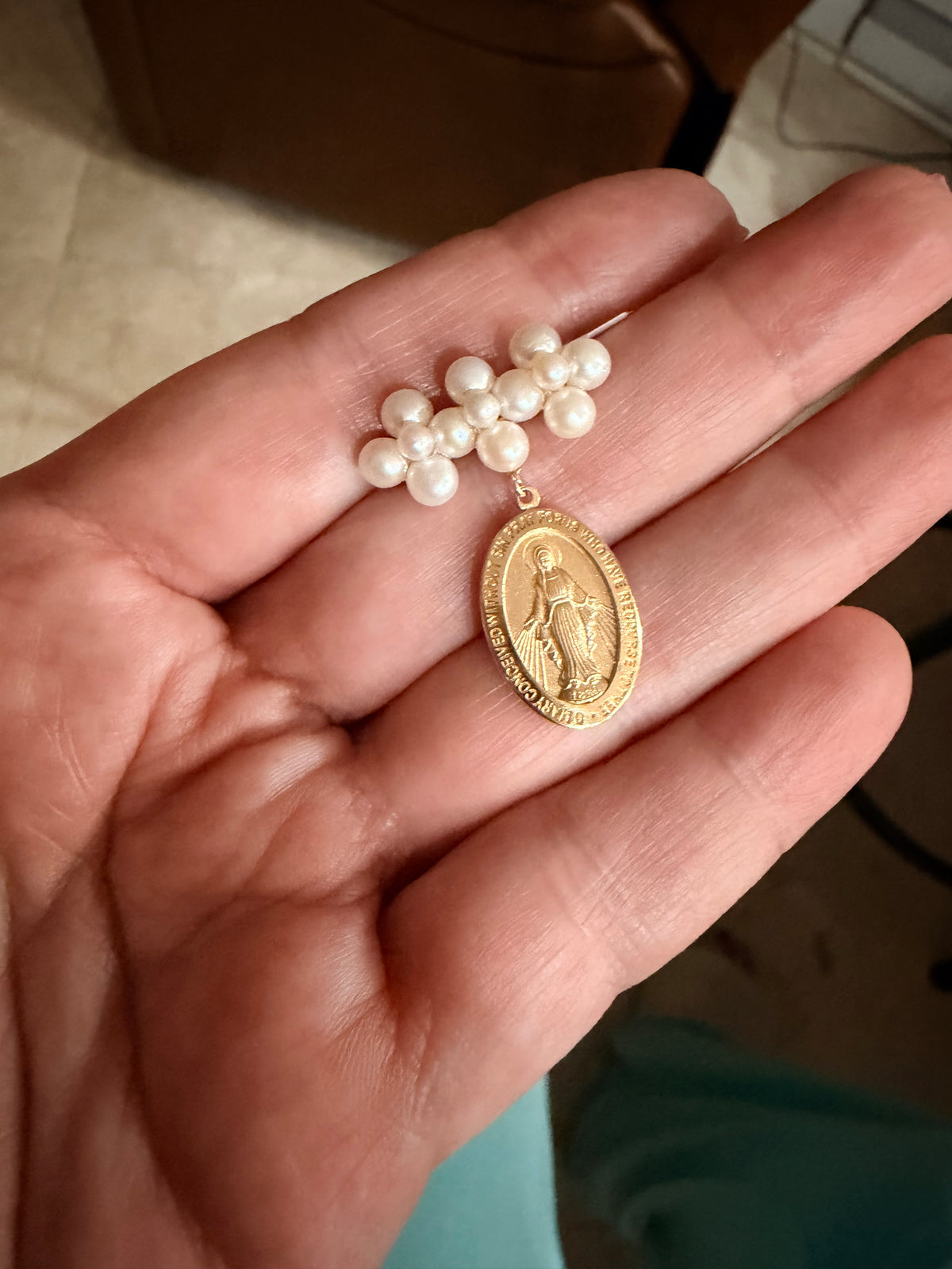 A close-up of a hand holding a pearl and gold religious pendant. The jewelry features a cluster of white pearls at the top and a gold oval medallion with a religious figure, likely Our Lady of Lourdes, hanging below.