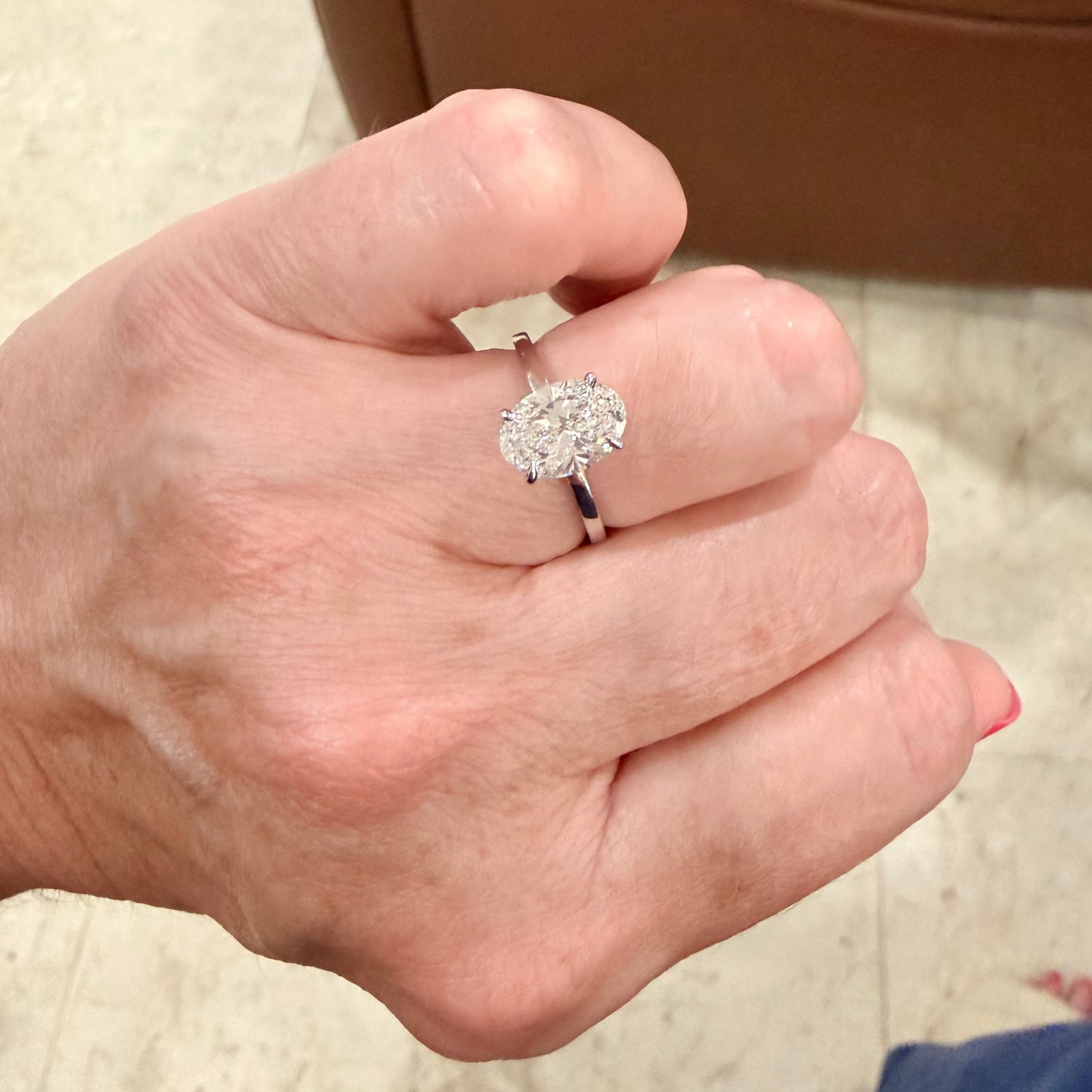 A close-up of a hand wearing a white gold engagement ring with an oval-cut diamond gemstone, photographed in an indoor setting with a brown leather chair and light tile floor in the background.