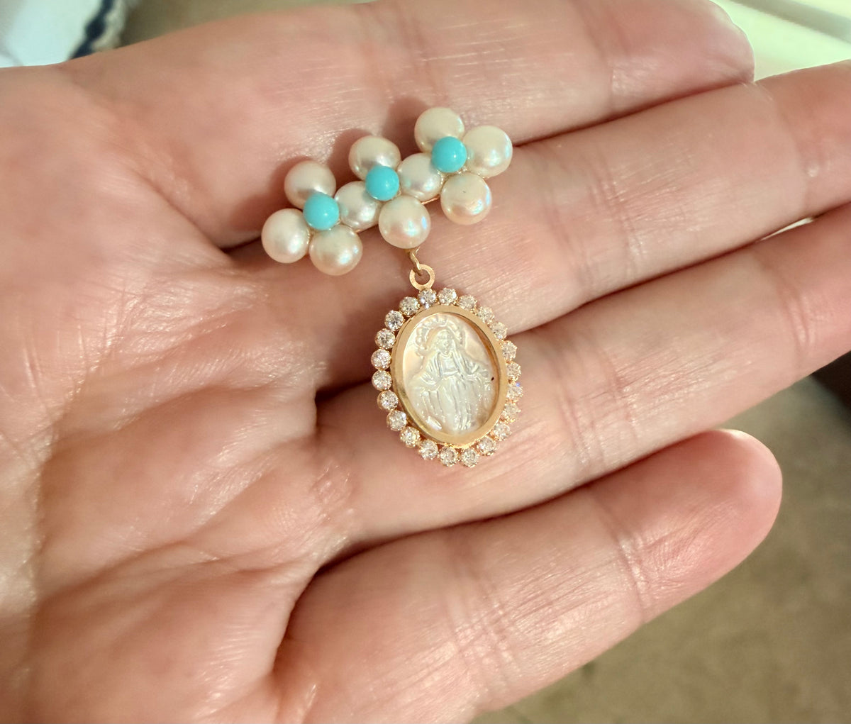 A close-up of a hand holding a pearl and turquoise brooch with a mother of pearl Miraculous medal pendant surrounded by cubic zirconia clear stones.