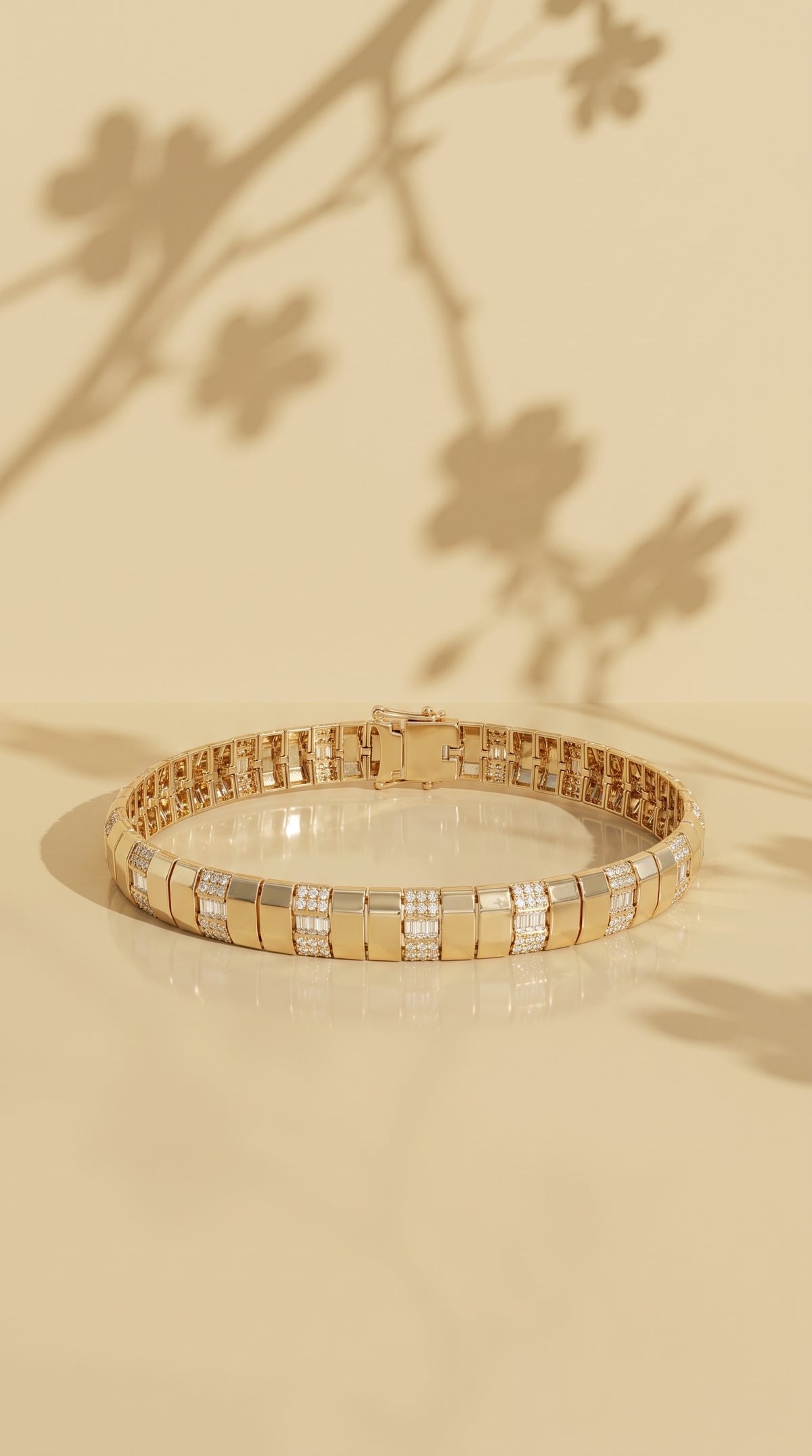 A 14k yellow gold contemporary tennis bracelet with rectangular links and rows of small channel set diamond baguettes and pave set round diamond stones, displayed on a warm beige reflective surface with soft floral shadows. 