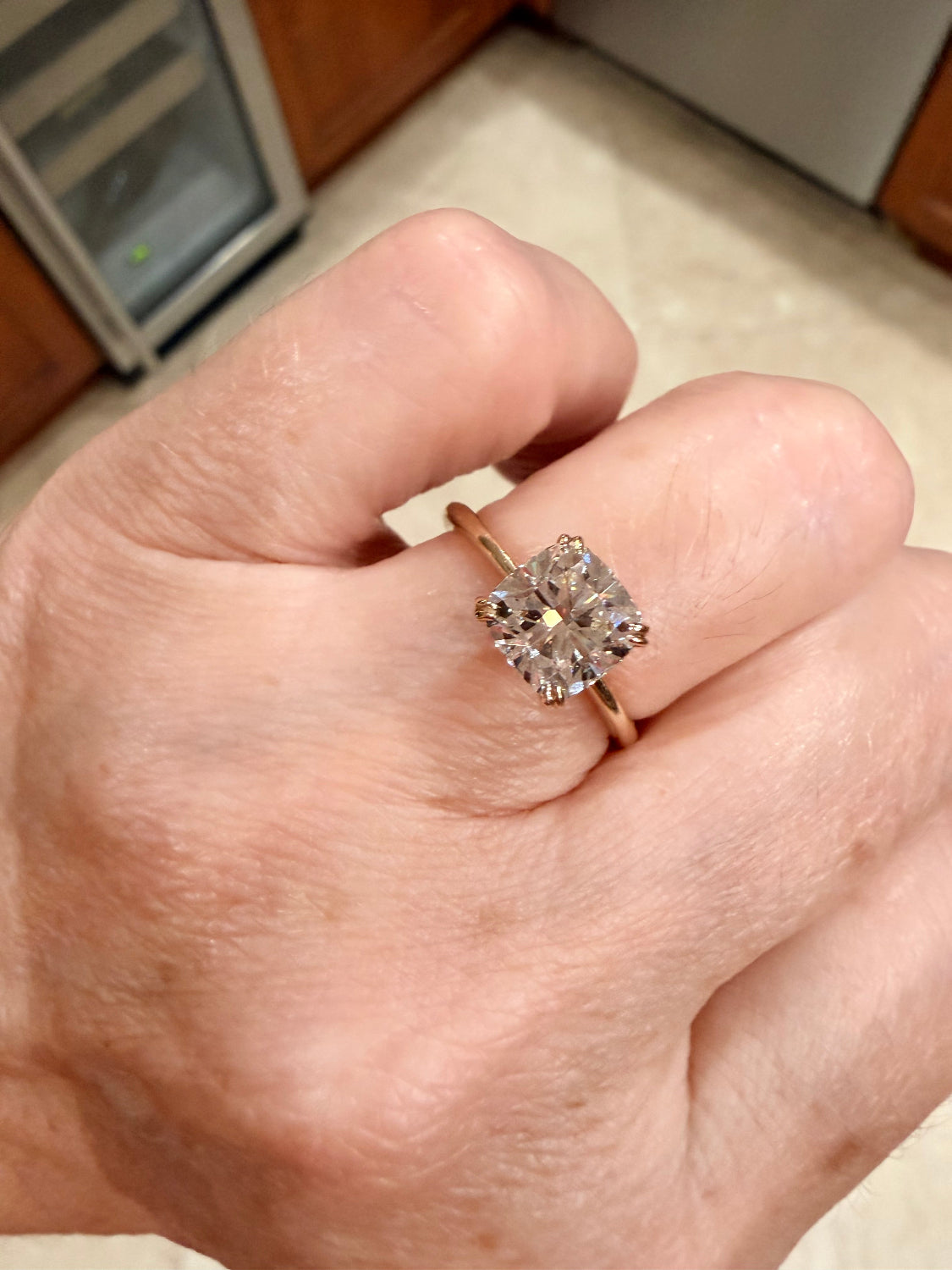 A close-up of a hand wearing a yellow gold ring with a large cushion-cut diamond gemstone, set against a blurred kitchen background.