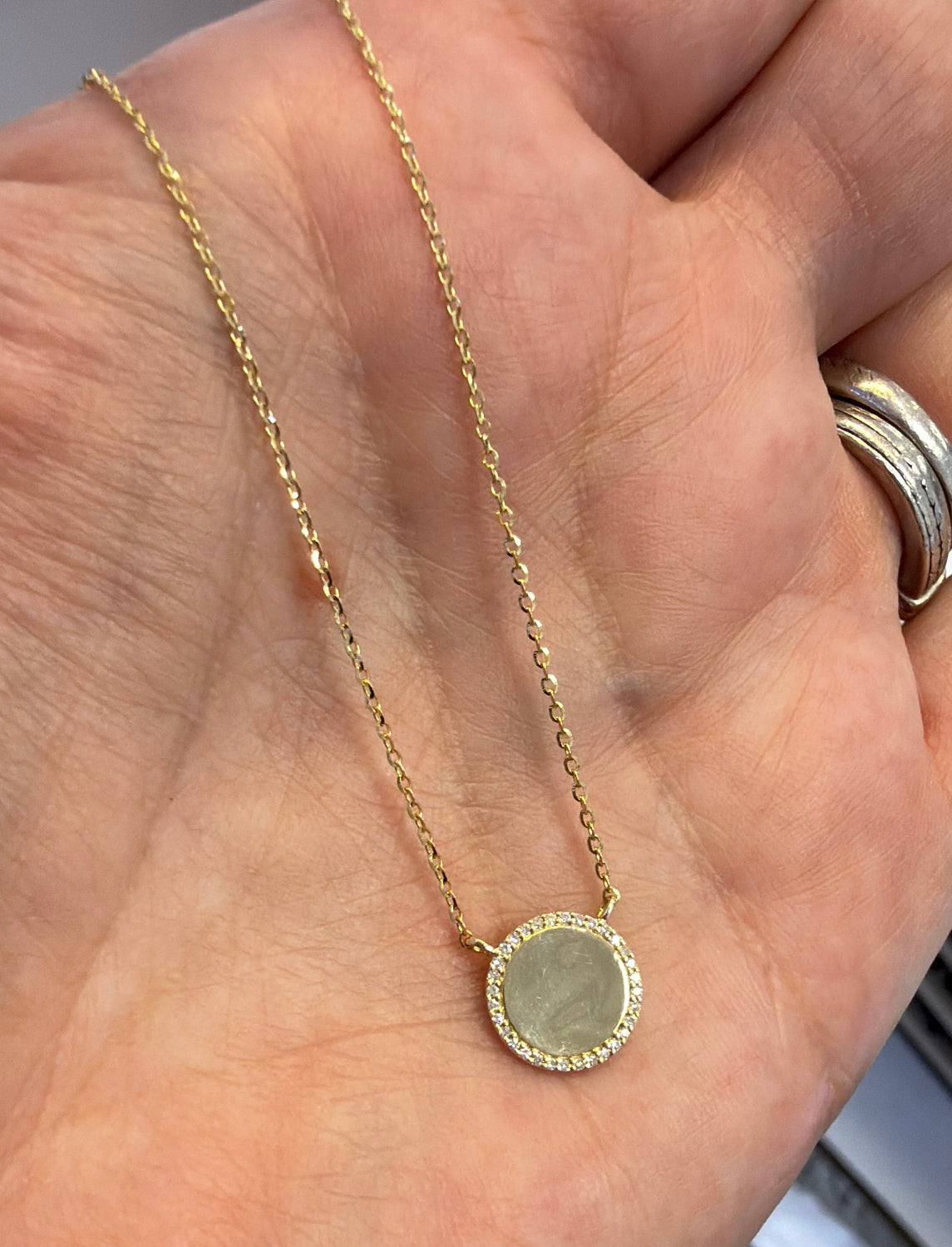 A delicate gold chain necklace with a round stone pendant, held in a person's hand. The pendant has a diamond pave border.
