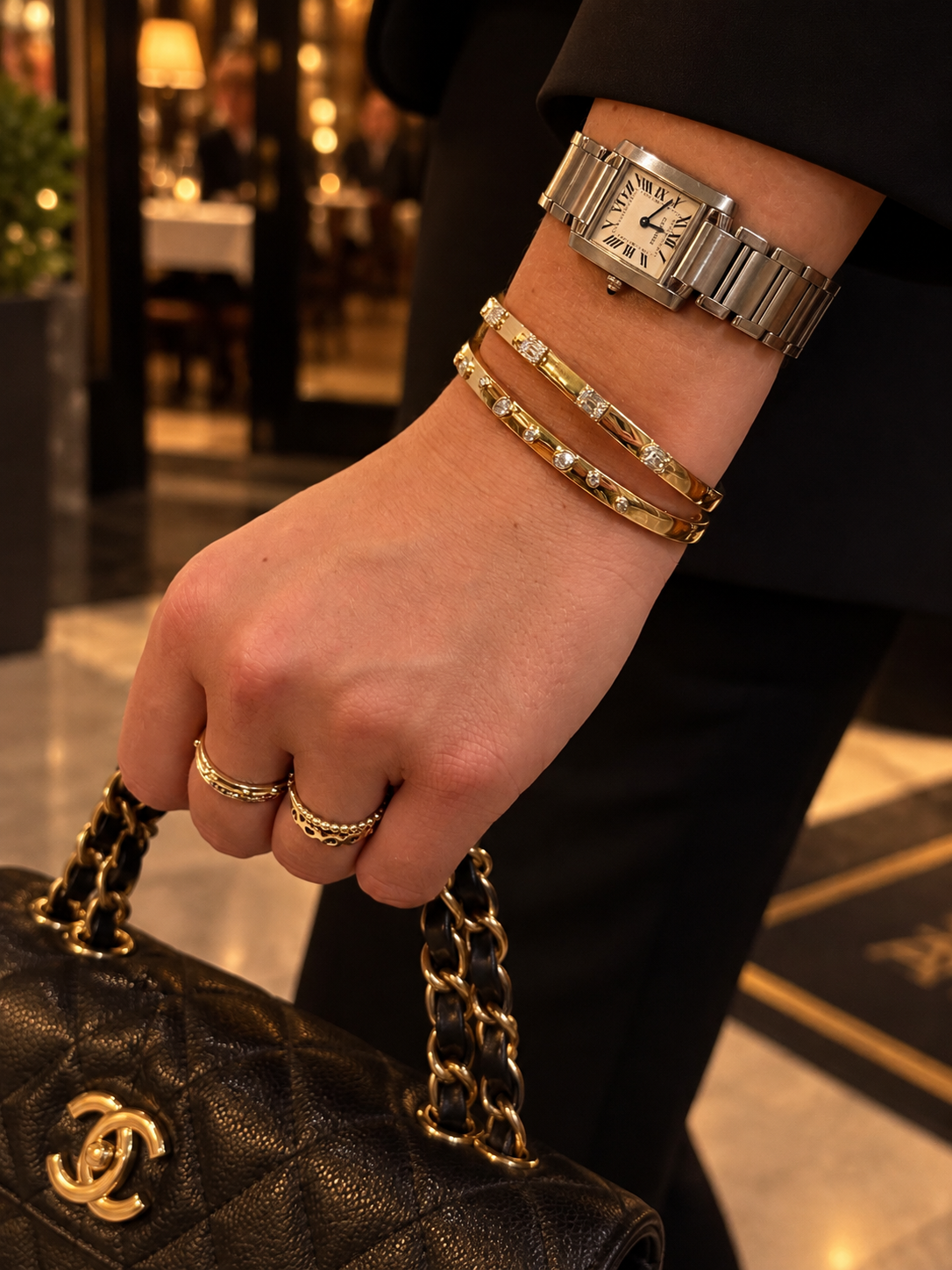 A close-up of a person's hand holding a black quilted Chanel handbag with gold chain straps, wearing a stainless steel Cartier watch and two 14K gold diamond bangle bracelets and rings, in a warm, upscale indoor setting.