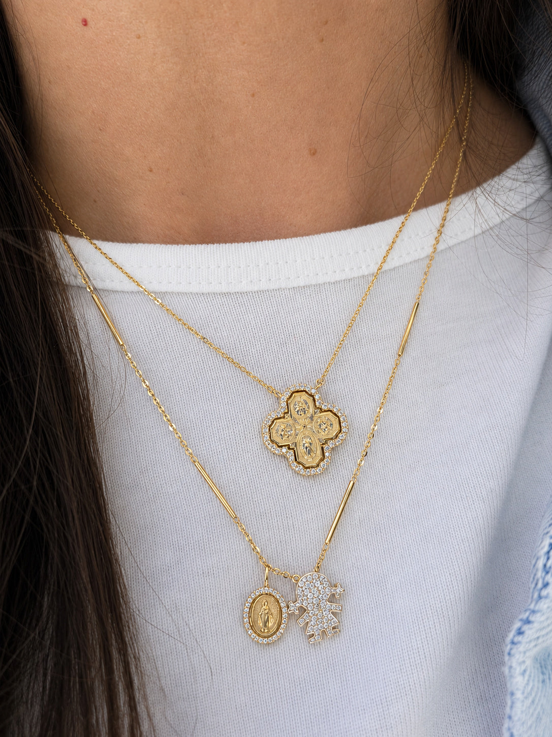 A close-up of a person wearing layered gold necklaces over a white t-shirt. The necklaces feature a gold 4-way cross pendant with diamonds on one chain and a longer chain with a small oval Miraculous  Medal medallion and a small diamonds and a pendant shaped like a girl-charm encrusted with diamond pave.