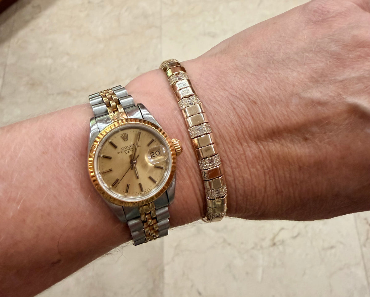 A close-up of a person's wrist wearing a gold-tone and stainless steel tone Rolex Datejust watch alongside a 14K gold contemporary wide diamond tennis bracelet.