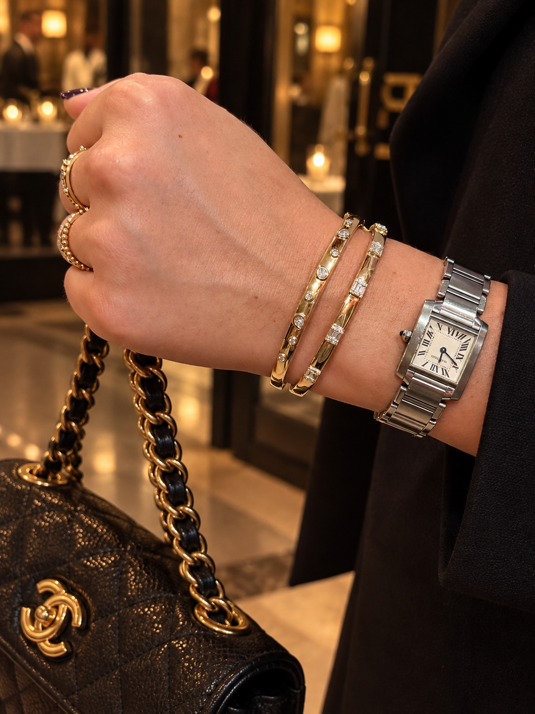 A close-up of a person's hand and wrist wearing multiple luxury accessories, including a stainless steel Cartier Tank watch, two 14K  gold diamond bangle bracelets, and gold rings, while holding a black quilted Chanel handbag with gold chain detailing.