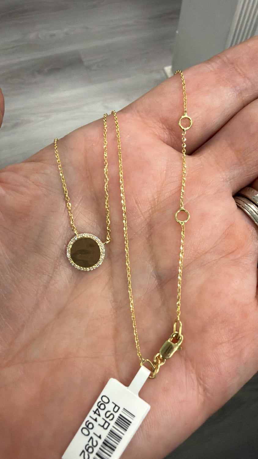 A gold necklace with a round pave  diamond halo high polish disc pendant is held in a person's hand. The necklace has a delicate chain with small circular links and a lobster clasp, and a white product tag is attached near the clasp.