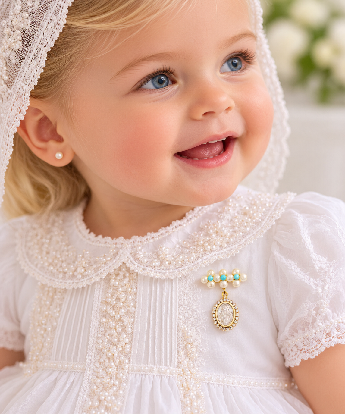 A close-up of a smiling young child wearing a white pearl-embellished dress with lace trim, a matching veil, and a small gold brooch with turquoise and pearl accents with a dangling mother of pearl Miraculous medal with cubic zirconia halo.