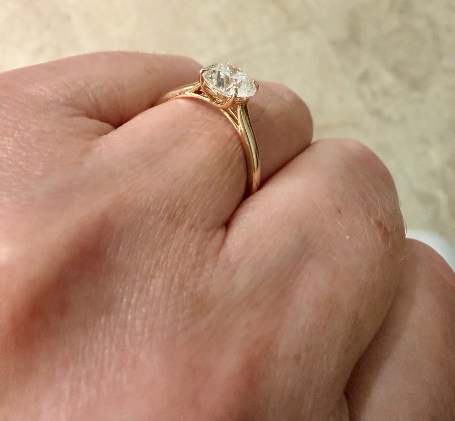 A close-up of a hand wearing a yellow gold engagement ring with a round-cut diamond gemstone in a prong setting, set against a neutral beige background.
