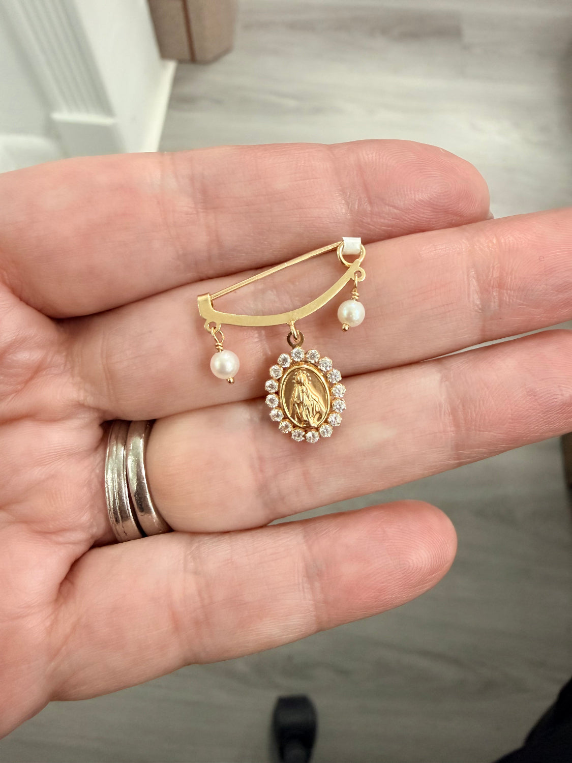 A gold and pearl religious brooch featuring a small oval Miraculous Medal pendant, held in a person's hand.