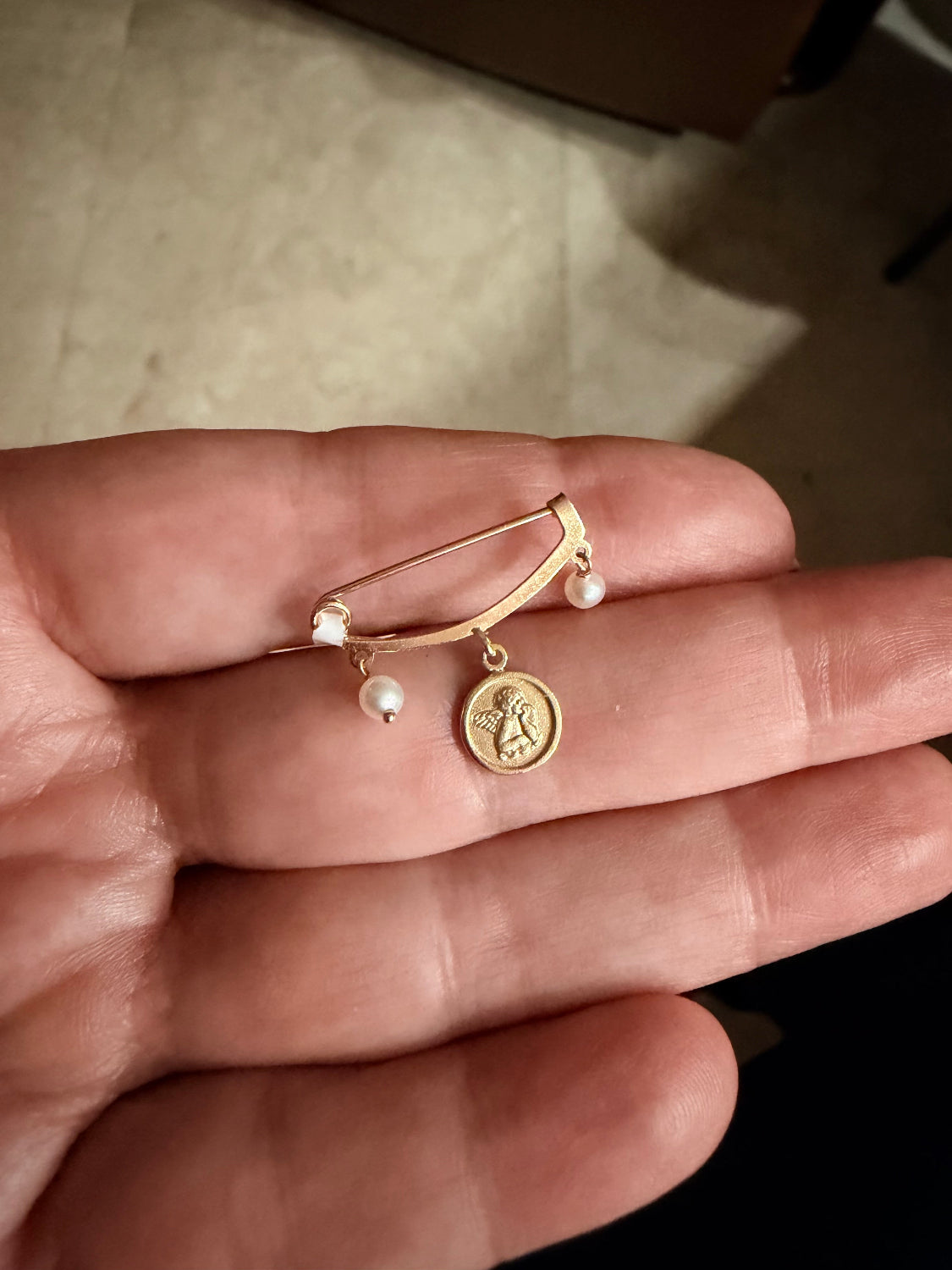 A gold baby pin brooch with two small white pearl and a round Angel medal charm, held in a person's hand.