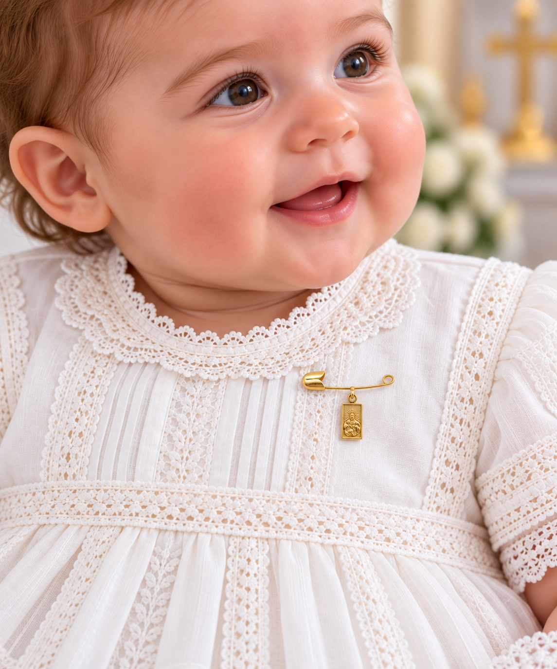 A close-up of a smiling baby wearing a white lace dress and a small gold baby keepsake pin with a hanging scapular medal charm, set against a softly blurred church background.
