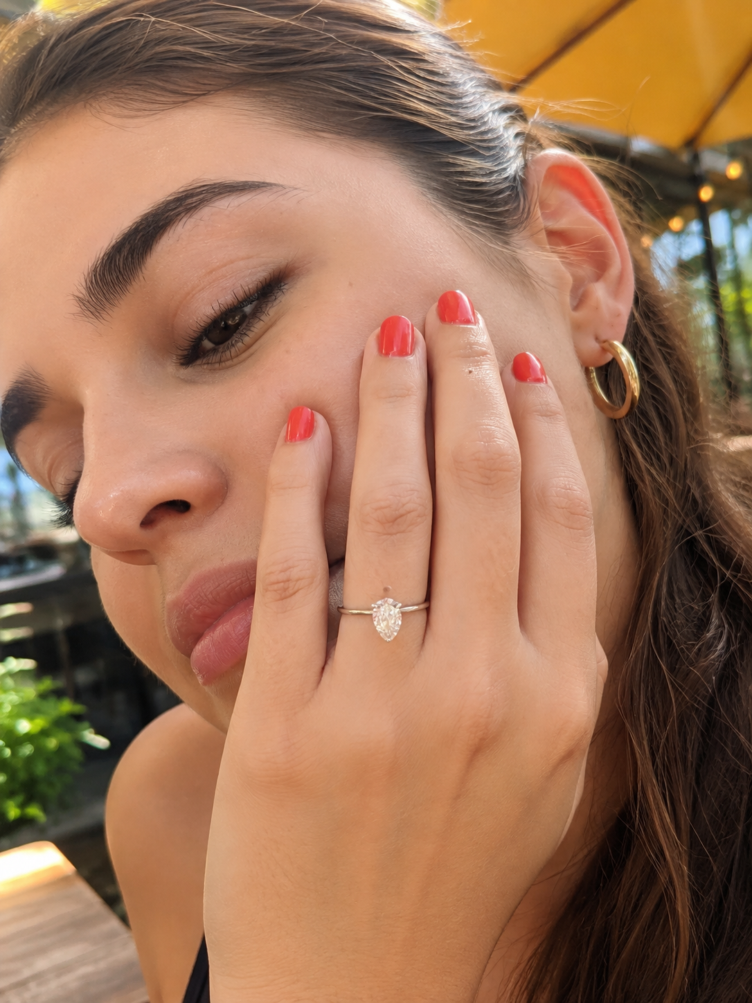 A close-up lifestyle shot of a woman wearing a white gold engagement ring with a pear shape-cut diamond gemstone on her ring finger. She has red nail polish and is wearing a gold hoop earring in an outdoor setting.