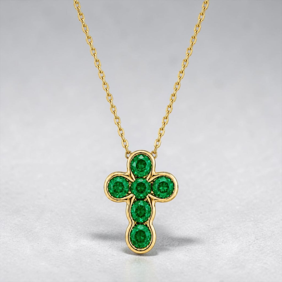 A gold cross pendant necklace featuring six round-cut green emerald gemstones, displayed on a soft gray background.