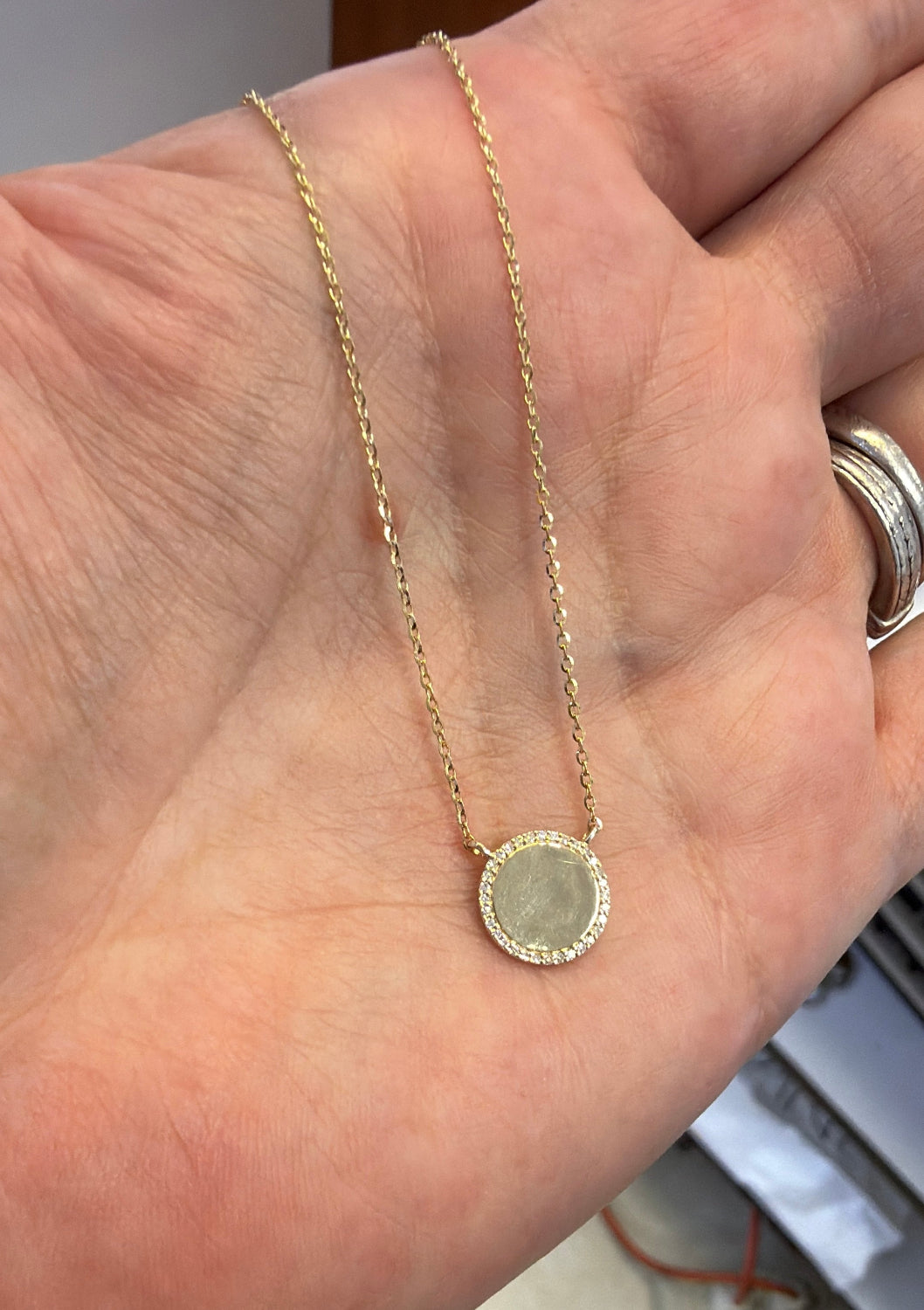 A delicate gold chain necklace with a round disc with diamond pendant, held in a person's hand.