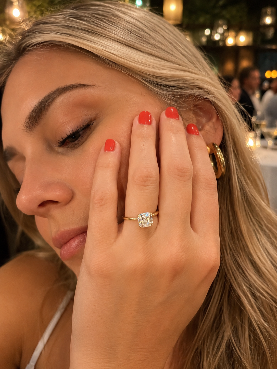 A close-up lifestyle shot of a woman wearing a yellow gold engagement ring with a cushion-cut diamond on her ring finger. She has blonde hair, red nail polish, and gold hoop earrings, with a softly blurred restaurant background.