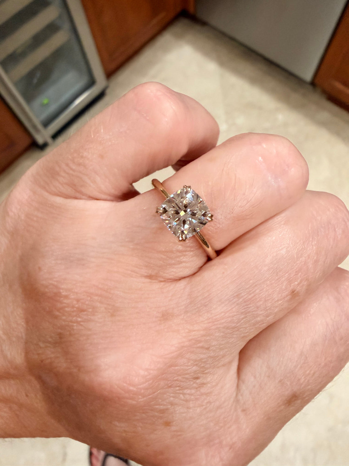 A close-up of a hand wearing a yellow gold ring with a cushion-cut diamond gemstone in a four-prong setting, with a kitchen background visible.