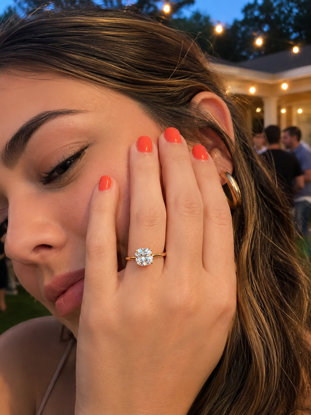 A close-up lifestyle shot of a woman wearing a yellow gold solitaire engagement ring with a cushion-cut diamond gemstone on her finger, with her hand resting against her cheek. The background shows an outdoor evening event with warm string lights and blurred people.