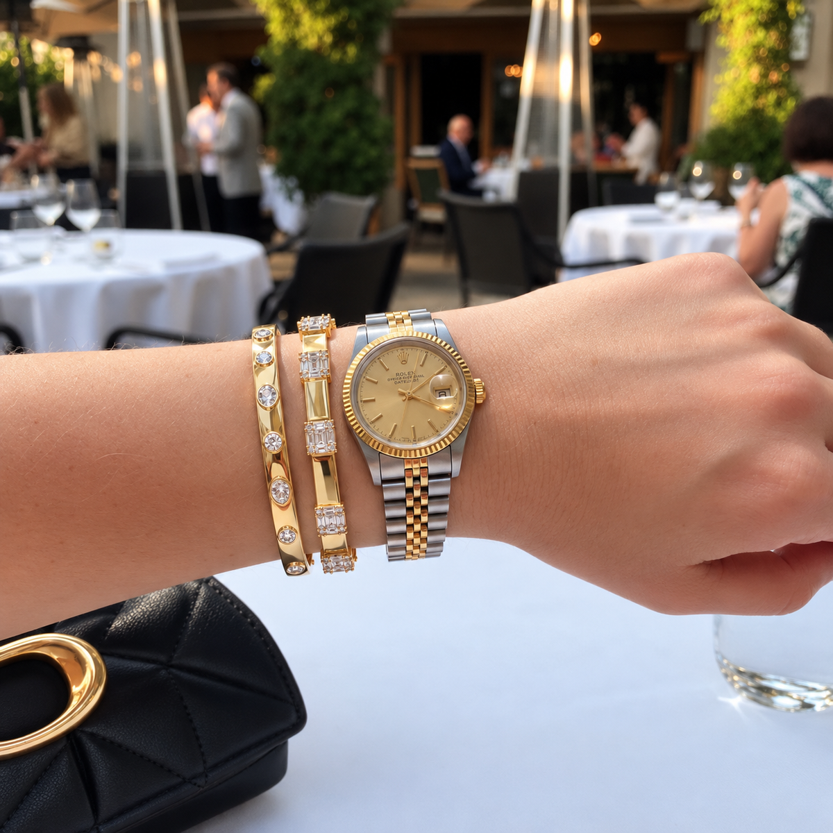 A close-up of a person's wrist wearing a gold-2-tone Rolex Lady Datejust watch with a two-tone steel and gold bracelet, alongside two gold bangles with diamonds, set against an outdoor restaurant background.