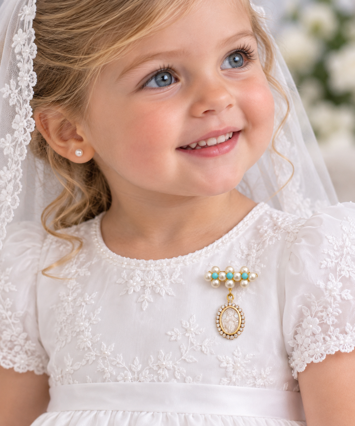 A young girl with blonde hair wearing a white lace dress, pearl stud earring, and a gold pendant brooch with turquoise and pearl accents with a dangling oval mother of pearl Miraculous medal with cubic zirconia halo , smiling in a softly blurred indoor setting.