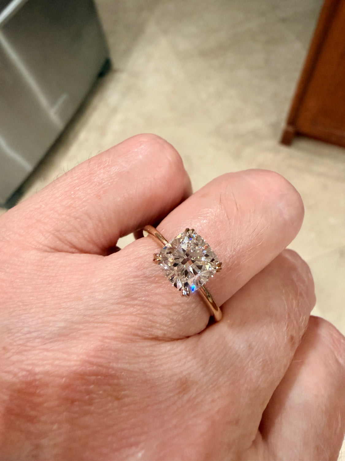 A close-up of a hand wearing a yellow gold ring with a cushion-cut diamond gemstone, set against a blurred indoor background.