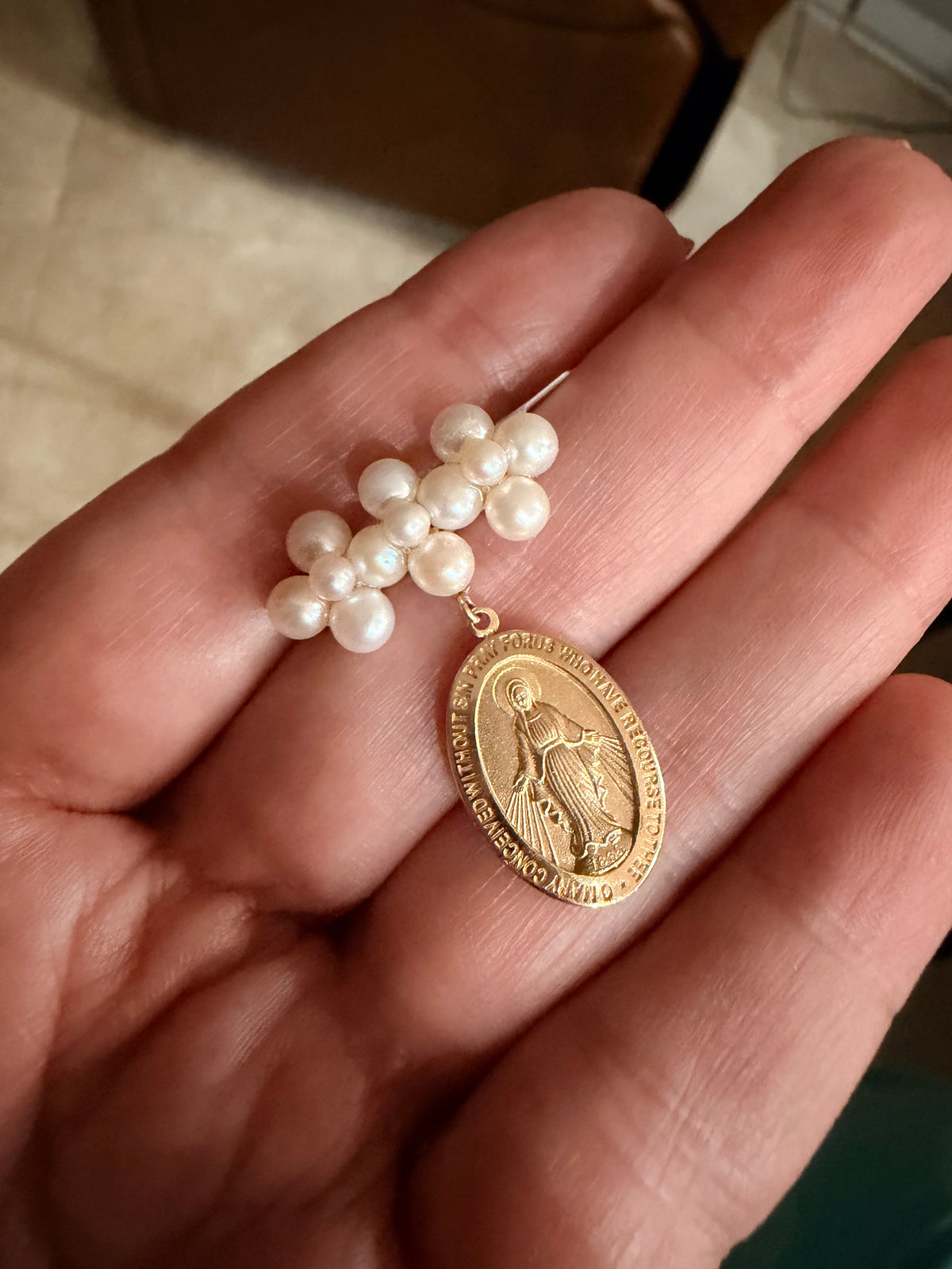 A close-up of a hand holding a pearl and gold religious Miraculous Medal pendant. brooch with pearls. The pendant features a gold oval medal with a figure of the Virgin Mary, suspended from a cluster of white pearl beads.