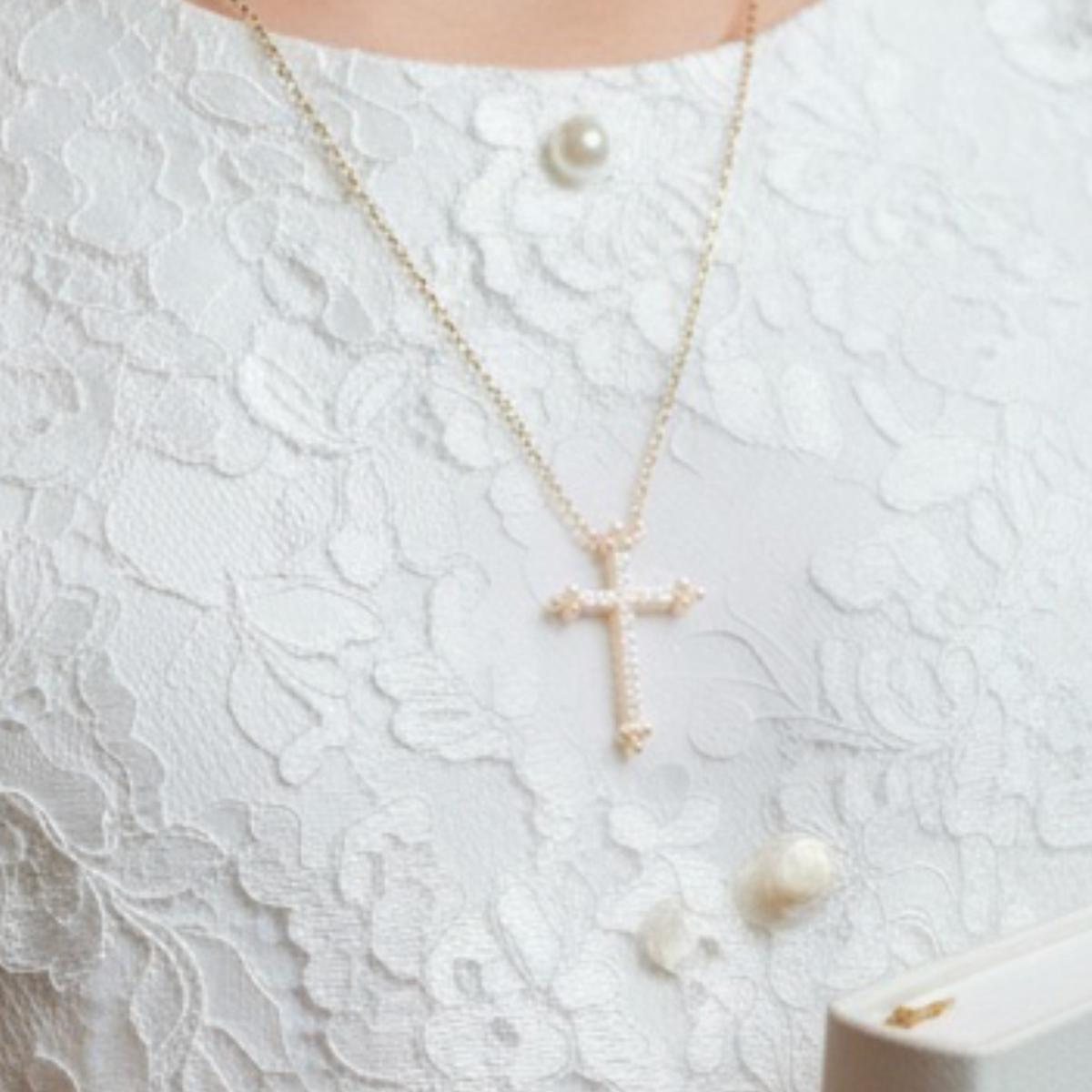 A close-up lifestyle shot of a woman wearing a delicate gold cross pendant necklace with pearl-like beads, styled with a white lace top and holding a white book.