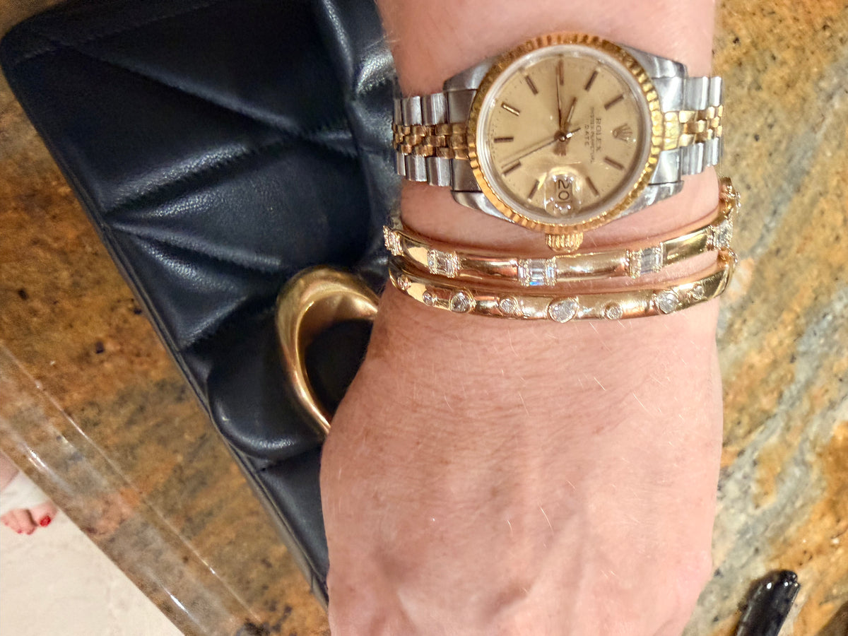 A close-up of a person's wrist wearing a two-tone gold and stainless steel Rolex Datejust watch, layered with two gold  diamond bangles, resting on a black quilted leather clutch over a granite countertop.