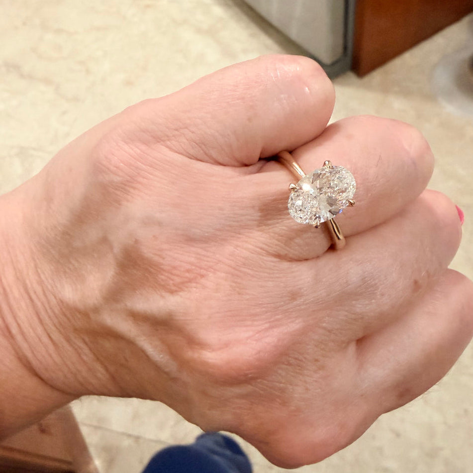 A close-up of a person's hand wearing a yellow gold solitaire engagement ring with a 3 carat oval cut diamond gemstone, set against a household background.