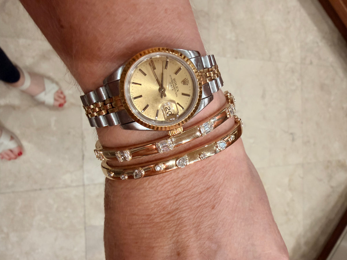 A gold and stainless steel two-tone Rolex watch with a gold dial and date window, worn on a wrist alongside two 14K  gold diamond bangles, against a beige tiled floor background.