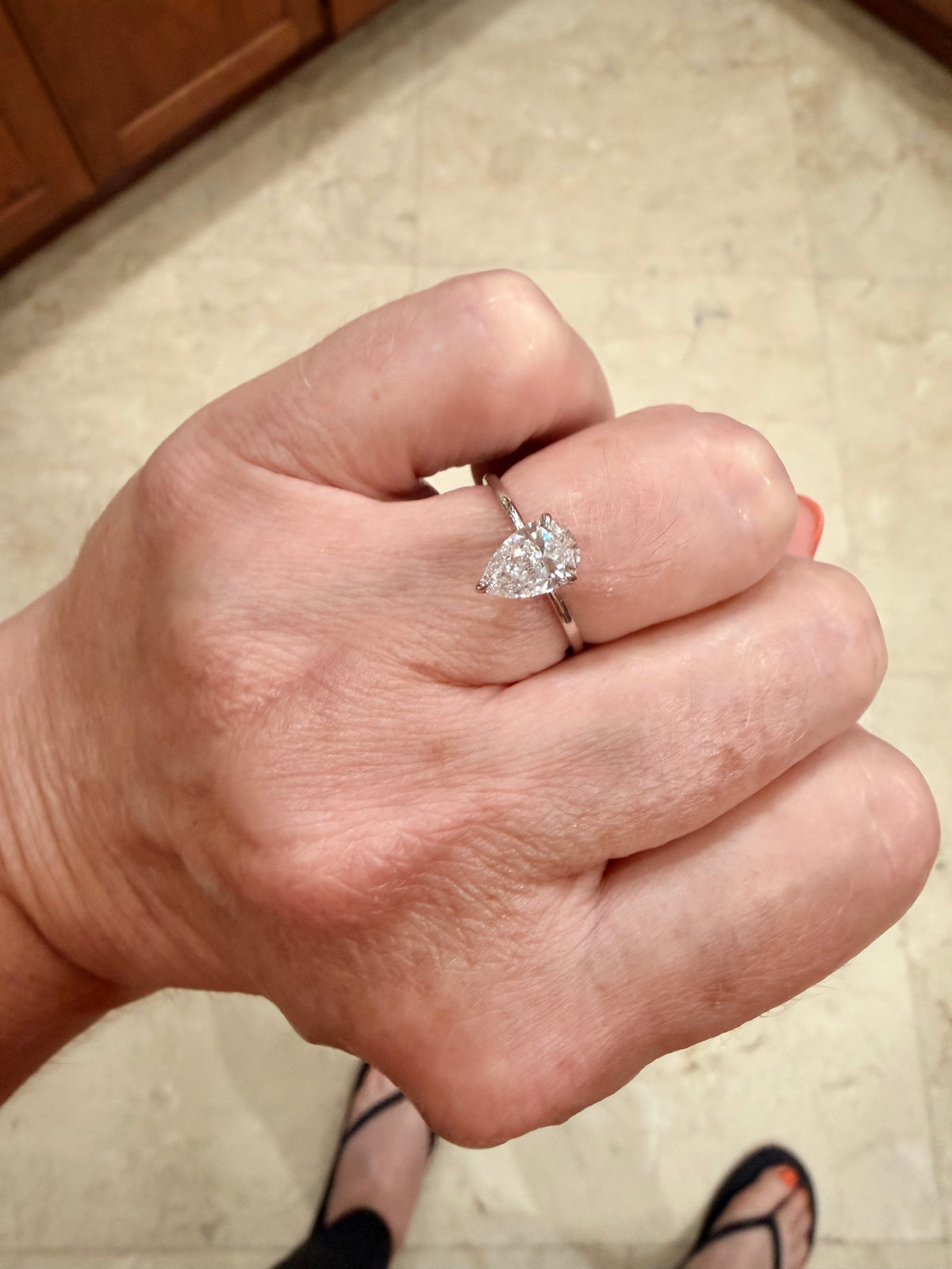 A close-up of a hand wearing a white gold engagement ring with a pear-cut clear diamond gemstone, set against a tiled floor background.